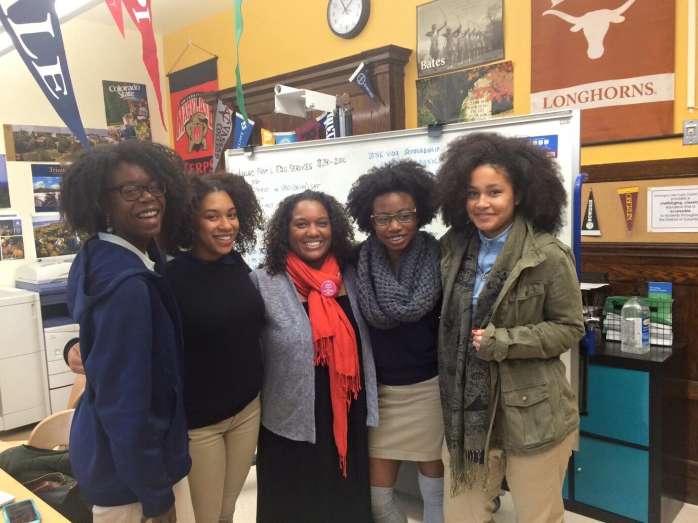 Five women smiling together indoors, showcasing diverse hairstyles and warm clothing.